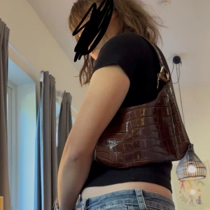 Brun axelväska i croco-imitation - Snygg brun axelväska i glansigt material med croco-mönster. Väskan har en klassisk bågform och stängs med dragkedja upptill. Perfekt storlek för det viktigaste och har guldfärgade metalldetaljer på remmen.