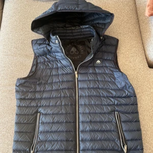 Mörk blå dunväst från Moose Knuckles - Snygg svart dunväst från Moose Knuckles med quiltad design och avtagbar huva. Västen har två dragkedjeförsedda fickor och tydlig logga på bröstet. Tillverkad i lätt, återvunnet material och fylld med dun. Perfekt för lager-på-lager-stil.