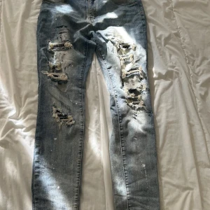 Ljusblå slitna skinny jeans med hål - Säljer ett par ljusblå slim jeans med rejält slitna detaljer och stora hål framtill. Jeansen har stänk av vitt och en cool, trasig look. Perfekta för dig som gillar streetstyle och vill sticka ut med din outfit.