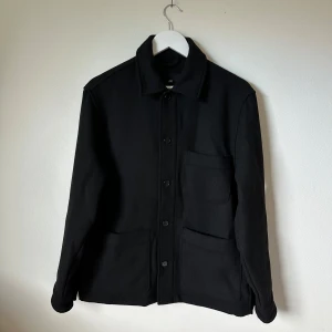 Svart overshirt från H&M i ullmix - Svart overshirt från H&M med klassisk krage och tre stora fickor framtill. Jackan har knäppning med knappar och är tillverkad i en mjuk ull- och polyestermix. Perfekt för lager-på-lager och enkel att styla till allt. 3U12