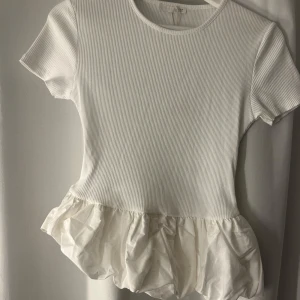 Vit ribbad topp med volangkant Zara - Supersöt vit topp från Zara med ribbad struktur och korta ärmar. Toppen har en rund halsringning och en volangig, ballongformad nederkant i ett glansigt tyg som ger en extra touch. Perfekt för dig som gillar en lekfull och trendig stil.