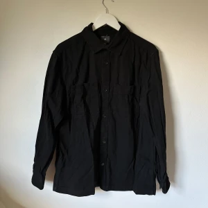 Svart överskjorta från H&M i bomull - Svart överskjorta från H&M i 100% bomull med klassisk krage, knäppning framtill och två bröstfickor. Långärmad modell med enkel, clean look som funkar till många stilar. Perfekt för dig som gillar minimalistiskt och stilrent. 3X12