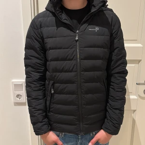 Svart pufferjacka från Pelle P - Säljer en svart pufferjacka från Pelle P med huva och dragkedja framtill. Jackan har quiltad design, två fickor med dragkedja och logga på bröstet samt ärmen. Perfekt för kalla dagar och riktigt snygg till jeans. Nypris 3500kr