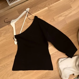 Svart oneshoulder topp - Säljer en svart oneshoulder topp med lång ärm på ena sidan och utan på andra. Enkel och stilren design, perfekt att matcha med jeans eller kjol. Materialet känns mjukt och stretchigt.