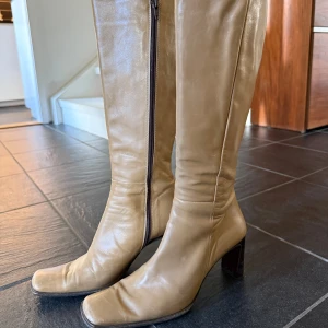 Beiga höga stövlar med klack - Snygga höga stövlar i beige, äkta skinn med fyrkantig tå och markerad klack. Dragkedja på insidan för enkel påtagning. Perfekta för dig som vill ha en stilren och trendig look med retrovibbar. 