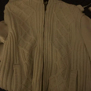 Vit stickad hoodie med dragkedja - Säljer en vit stickad hoodie med dragkedja och fickor framtill. Tröjan har ett snyggt kabelstickat mönster på framsidan och ribbade detaljer på baksidan. Insidan av huvan har ett camouflagemönster som ger en cool kontrast.