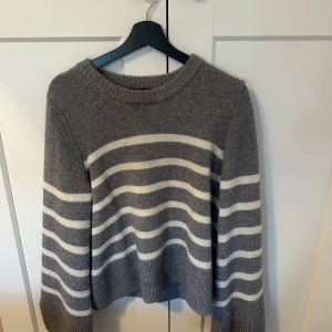 Gråvit randig stickad tröja Wakakuu icons  - Mysig stickad tröja i grått med vita horisontella ränder. Tröjan har rund halsringning, långa ärmar och ribbade muddar. Perfekt för kyliga dagar och enkel att matcha med jeans eller kjol. 35% Kashmir 65% Ull 