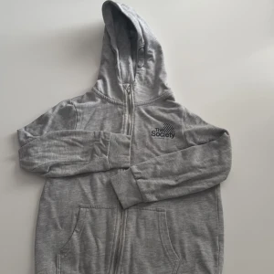 Grå hoodie från 157 med dragkedja - Grå hoodie från 157 med svart The Society-tryck på bröstet. Tröjan har huva, hel dragkedja framtill och två stora fickor. Tillverkad i mjuk bomullsmix och har långa ärmar med muddar. Perfekt för chill dagar eller när du vill vara extra bekväm.