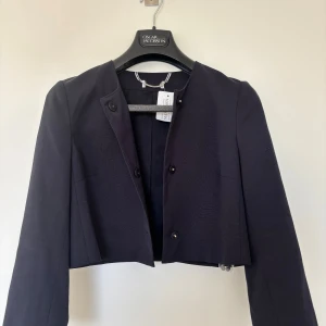 Mörkblå croppad kavaj från Oscar Jacobson - Säljer en stilren mörkblå croppad kavaj från Max Mara. Jackan har rund halsringning, raka axlar och knäppning framtill med tre knappar. Perfekt för dig som gillar en minimalistisk och modern look. Materialet känns mjukt och har en lätt struktur. 
