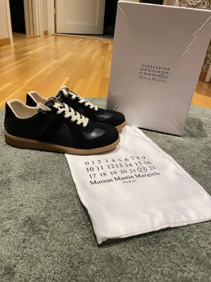 Maison Margiela svarta sneakers - Maison Margiela sneakers i svart skinn och mocka med beige snörning och gummisula. Klassisk låg modell med rund tå och platt sula. Märkets logga syns på sulan och medföljande dustbag. Perfekt för dig som gillar stilrena och exklusiva sneakers.