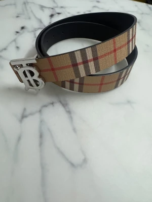 Burberry bälte  - Säljer ett snyggt Burberry bälte med klassiskt rutig design i beige, brun, svart och röd. Bältet har ett stilrent silverfärgat B-spänne i metall och är tillverkat i skinn med textil på ovansidan. Perfekt för att lyfta din outfit med en ikonisk touch.