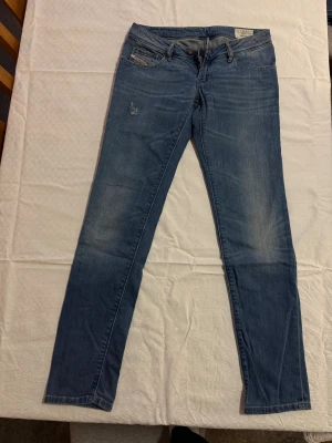 Blå Diesel NEVY jeans W29 L32 - Snygga blå jeans från Diesel, modell NEVY med stretch. Klassisk femficksdesign, diskret Diesel Industry-detalj på myntfickan och lagad detalj på låret. Smal passform och raka ben, perfekt till sneakers eller boots.