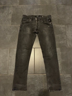 Svarta Replay jeans slim fit - Svarta jeans från Replay med klassisk femficksdesign och diskreta slitningar. Jeansen har smal passform och raka ben, med snygga detaljer på bakfickorna. Perfekta för dig som gillar en stilren och modern look.