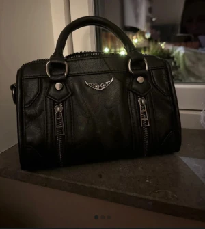 Liknande zadig Voltaire väska  - Cool svart handväska i skinnimitation med två rejäla handtag och snygga silverfärgade detaljer. Väskan har två dekorativa dragkedjor framtill och en liten metallvinge som detalj. Perfekt storlek för det viktigaste och ger en edgy vibe till din outfit. Inte en äkta zadig Voltaire utan en liknande. 