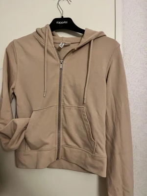 Beige hoodie med dragkedja från H&M - Snygg beige hoodie från H&M Basic i storlek S. Tröjan har huva med snörning, praktisk dragkedja framtill och en stor magficka. Tillverkad i mjuk bomullsmix och har långa ärmar. Perfekt för chill dagar och enkel att matcha med jeans eller leggings.