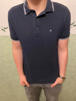 Mörkblå piké från Replay - Snygg mörkblå pikétröja från Replay med vit broderad logga på bröstet. Klassisk krage med vita ränder och tre knappar framtill. Materialet är mjuk bomull och passformen är normal. Perfekt för dig som vill ha en stilren och enkel look.