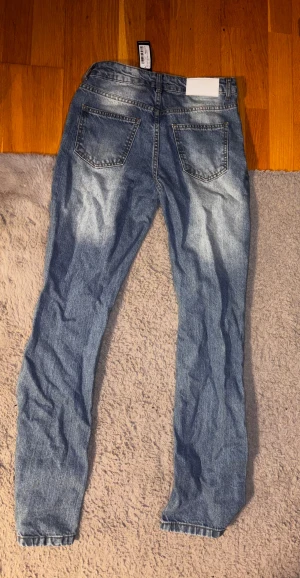 Blå jeans från BoohooMAN, storlek 28 - Snygga blå jeans från BoohooMAN i storlek 28. Modellen har klassisk femficksdesign, ljus tvätt med slitningar och raka ben. Jeansen har normal passform och är tillverkade i bomullsmaterial för skön känsla. Perfekt för dig som gillar en avslappnad stil. Helt oanvända.