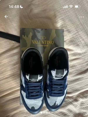 Valentino Garavani camo sneakers - Snygga Valentino Garavani sneakers med blå och vita camo-detaljer. Ovandel i mesh och mocka, svart och vit färgkombination, samt grov gummisula för extra grepp. Klassisk snörning och coola rockstuds på hälen. Perfekt för dig som gillar exklusiva streetwear-vibbar.