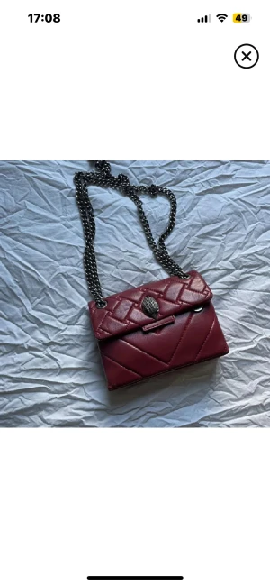 Mörkröd quiltad axelväska med kedja - Snygg mörkröd quiltad axelväska med silverfärgad kedja och coolt dödskalle-spänne framtill. Väskan har en stilren, rektangulär form och är tillverkad i skinnimitation. Perfekt för dig som gillar edgy detaljer och vill sticka ut.