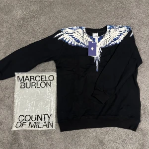 Marcelo Burlon svart tröja med vingar - Svart tröja från Marcelo Burlon County of Milan med ikoniskt vingtryck i vitt och blått över axlar och bröst. Rund halsringning och långa ärmar. Tillverkad i mjuk bomull för en skön känsla. Perfekt statement-piece för dig som vill sticka ut.