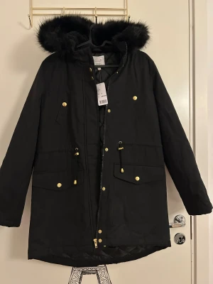 Svart parkas med fuskpäls från Club XPRS - Svart parkas med fuskpäls på huvan i storlek S från Club XPRS. Jackan har guldfärgade knappar, dragkedja och snörning i midjan. Insidan är quiltad för extra värme och har två stora fickor framtill. Jackan är i nytt skick, aldrig använd och har prislappen kvar. 