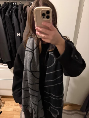 Svart och grå halsduk med grafiskt mönster - Snygg halsduk i svart och grått med ett coolt grafiskt mönster i kontrasterande färger. Halsduken är lång och bred, perfekt att svepa runt halsen för extra stilpoäng. Materialet är stickat och känns mjukt mot huden. Passar dig som gillar minimalistisk design och vill ha något som sticker ut.