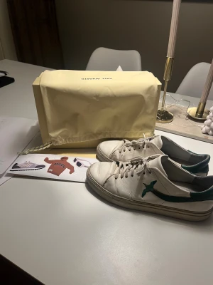 Axel Arigato vita sneakers med grön fågel - Säljer ett par vita sneakers från Axel Arigato med grön fågelbroderi på sidan och gröna detaljer vid hälen. Skorna har rund tå, snörning och platt sula. Tillverkade i skinn och levereras med originalkartong och dustbag. Perfekt för dig som gillar stilrena och coola sneakers.