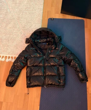 Ralph Lauren jacka - Säljer denna snygga svarta pufferjackan från polo Ralph lauren med glansig finish och klassisk röd logga på bröstet. Har knappt använt den och kvitto finns!