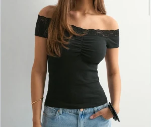 Svart offshoulder topp med spets - Snygg svart offshoulder topp med korta ärmar och spetsdetalj längs halsringningen. Toppen är ribbad och har en smickrande rynkning framtill. Perfekt för dig som gillar en stilren look med lite extra edge.