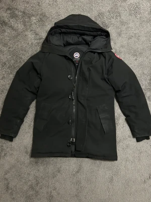 Svart jacka från Canada Goose - Svart jacka från Canada Goose. Har använts och är i helt okej skick. Den har lite slitage som blekningar och ärmarna är lite skadade(se bild 4). 