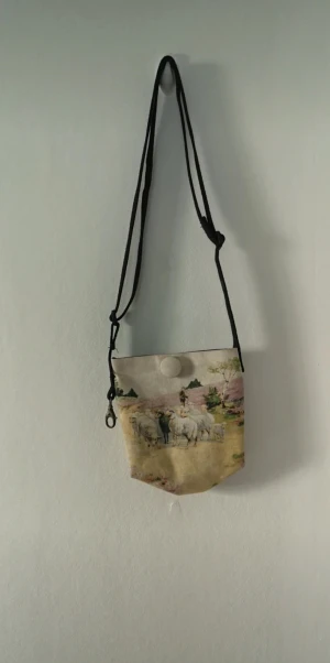 Beige axelväska med fårmotiv - Unik beige axelväska i bomull med ett konstnärligt tryck av får och landskap på framsidan. Väskan har svart justerbar axelrem, en stor rund knapp som stängning och en metallhake i sidan.