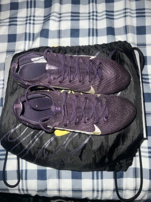 Nike Air Zoom Mercurial fotbollsskor lila - Säljer ett par Nike Air Zoom Mercurial fotbollsskor i mörklila med silverfärgade detaljer och gul sula. Skorna har snörning, platt sula med dobbar och KM-logga på sidan. Ovandelen är i syntetmaterial med stickad känsla för extra komfort och passform.