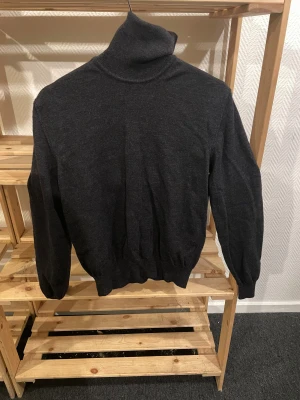 Mörkgrå polotröja Tiger of Sweden - Snygg mörkgrå polotröja från Tiger of Sweden i mjuk merinoull som ger en väldigt mysig känsla. Tröjan har lång ärm, ribbade muddar och klassisk hög polokrage. Perfekt för dig som gillar stilrena och tidlösa plagg med en chill vibe.