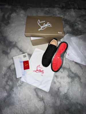 Svarta sneakers från Christian Louboutin - Snygga svarta sneakers från Christian Louboutin med klassisk rund tå och platt sula. Skorna har svart snörning och är tillverkade i mocka med beige innersula. Ikonisk röd undersida med Louboutin-logga. Perfekt för dig som gillar exklusiv streetstyle.