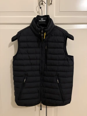 Svart dunväst från Parajumpers - Snygg svart dunväst från Parajumpers med quiltad design och hög krage. Västen har dragkedja framtill, två sidofickor med dragkedja och en gul detalj vid halsen. Insidan är grå och materialet känns lätt och smidigt. Perfekt för lager-på-lager under kyliga dagar och perfekt för att ha den under vår/höst/sommar. Kvitto finns och den är knappt använd