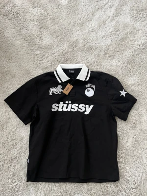 Svart Stüssy skjorta med tryck - Svart skjorta från Stüssy med vit kontrastkrage och stora vita tryck fram och bak. Coolt lejonbroderi, stjärna på ärmen och klassisk Stüssy-logga. Skjortan har knappar vid halsen och är tillverkad i mjuk bomull. Perfekt streetwear-vibe med World Tour-tryck på ryggen.