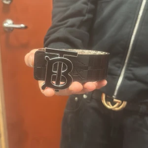 Burberry TB bälte med rutigt mönster - Säljer ett Burberry bälte med ikoniskt TB-spänne i silverfärgad metall. Ena sidan är svart med diskret rutigt mönster, andra sidan har klassiskt Burberry-rutigt i beige, svart och rött. Bältet är brett och tillverkat i skinn med lyxig känsla. Perfekt för att lyfta din outfit.