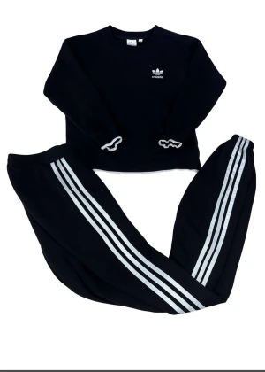 Adidas Set Tröja och Byxor Storlek S - Svart Adidas sweatshirt med klassisk vit treklöver-logga på bröstet. Tröjan har rund hals, långa ärmar och ribbade muddar med vit kant. Materialet är mjuk bomull och polyester, perfekt för chill eller träning. Enkel och stilren design som funkar till allt.