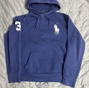 Blå hoodie från Polo Ralph Lauren - Snygg blå hoodie från Polo Ralph Lauren med stor vit broderad logga på bröstet och siffran 3 på ärmen. Tröjan har huva med snörning och en klassisk magficka. Perfekt för chill dagar och enkel att matcha med jeans.