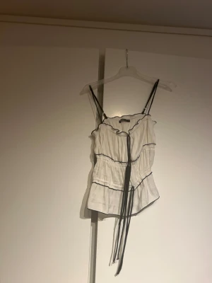  Vit linnetopp med svarta detaljer  - Säljer en vit linnetopp med svarta tunna axelband och svarta sömmar som ger en snygg kontrast. Toppen har volangdetaljer och snörning framtill, samt en asymmetrisk nederkant. Perfekt för dig som gillar en feminin och trendig look.