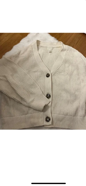 Beige stickad kofta från H&M - Snygg beige stickad kofta från H&M i storlek M. Koftan har grovstickat mönster, v-ringad hals och stora, coola knappar framtill. Perfekt att slänga över en t-shirt eller linne när det blir lite kyligt. Långärmad och med breda muddar för extra comfy vibe.