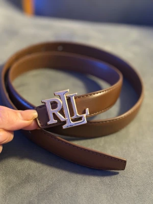 Brunt skärp från Ralph Lauren RL - Snyggt brunt bälte från Ralph Lauren med ett stort RL-spänne i guldmetall. Bältet är gjort i slätt skinn och har klassisk design med flera hål för justering. Perfekt accessoar för att lyfta din outfit med en stilren touch.