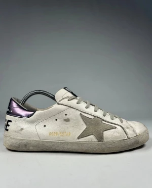 Golden Goose vita sneakers med stjärna - Golden Goose sneakers i vitt skinn med grå mockadetaljer och en stor stjärna på sidan. Skorna har grå snörning, platt sula och metallicfärgad häl. GGDB-logga på plösen och guldtext på sidan. Perfekt för dig som gillar en cool och avslappnad stil. Skriv om ni har frågor eller funderingar!😊