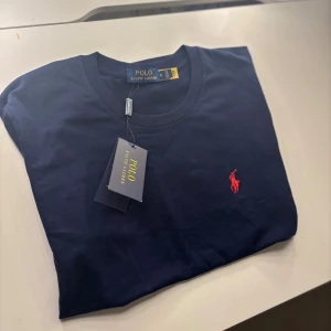Mörkblå t-shirt från Polo Ralph Lauren - Snygg mörkblå t-shirt från Polo Ralph Lauren i mjuk bomull. Klassisk rund hals och korta ärmar. På bröstet finns det ikoniska röda broderade Polo-logot. Perfekt för dig som gillar stilrena och tidlösa plagg med premiumkänsla.