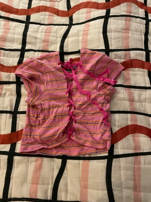 Randig rosa croppad topp med snörning - Säljer en söt croppad topp i rosa med lila och gula ränder, korta ärmar och snörning baktill med breda rosa band. Framsidan har tryck med två lila Care Bears och grönt kantband vid halsen. Perfekt för dig som gillar färgglada och lekfulla plagg.