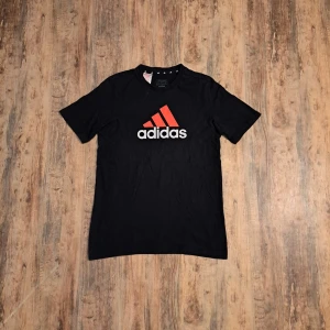 Svart Adidas t-shirt med röd logga - Svart t-shirt från Adidas med stor röd logga och vit text på bröstet. Klassisk rund hals och korta ärmar. Materialet är mjuk bomull som känns skönt mot huden. På ryggen finns en mindre röd Adidas-logga tryckt upptill.