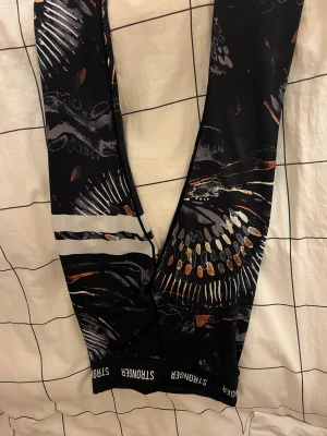 Stronger Safari leggings med mönster - Snygga leggings från Stronger med mönster i orange, grått och vitt. Vita ränder på ena benet. De är använda några gånger och blivit lite utböjda men annars i bra skick. Perfekta för träning eller chill. Materialet är stretchigt syntet som andas. 
