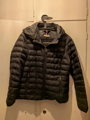 Svart dunjacka Tommy Hilfiger XL - Svart dunjacka från Tommy Hilfiger i storlek XL. Jackan har huva, två sidofickor med dragkedja och quiltad design. Klassisk logga på bröstet och snygga detaljer med röd och vit rand på insidan av kragen. Perfekt för kalla dagar.
