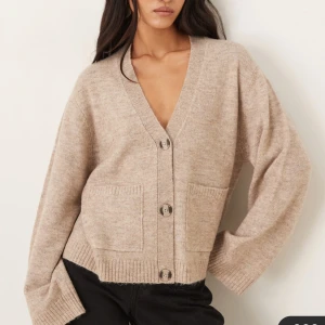 Beige stickad kofta med knappar - Mysig beige stickad kofta med v-ringning och stora knappar framtill. Koftan har två rejäla fickor och ribbade muddar vid ärmslut och nederkant. Passformen är loose och ärmarna är långärmade, perfekt för att slänga på sig när det är lite kyligt.