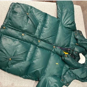Grön pufferjacka från Polo Ralph Lauren - Snygg grön pufferjacka från Polo Ralph Lauren med gul broderad logga på bröstet. Jackan har huva, dragkedja och knappar framtill, samt två stora fickor med knappstängning. Perfekt för kalla dagar och har en cool, sportig vibe.